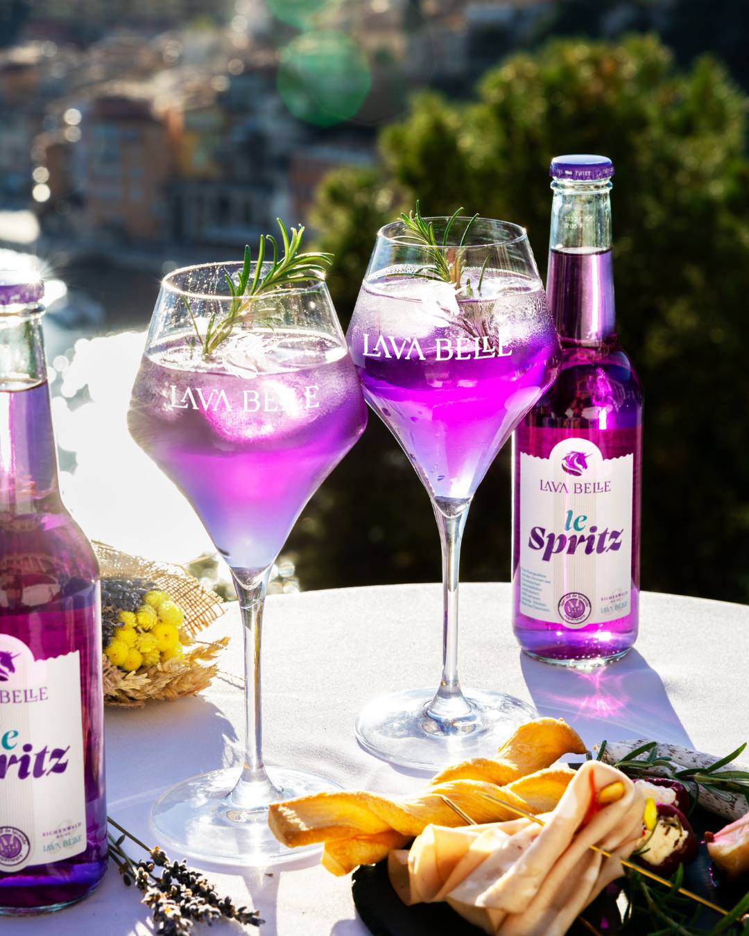Zwei le Spritz Flaschen und Lav'a Belle Gläser mit Rosmarin auf gedecktem Tisch mit Snacks über einem Küstenort