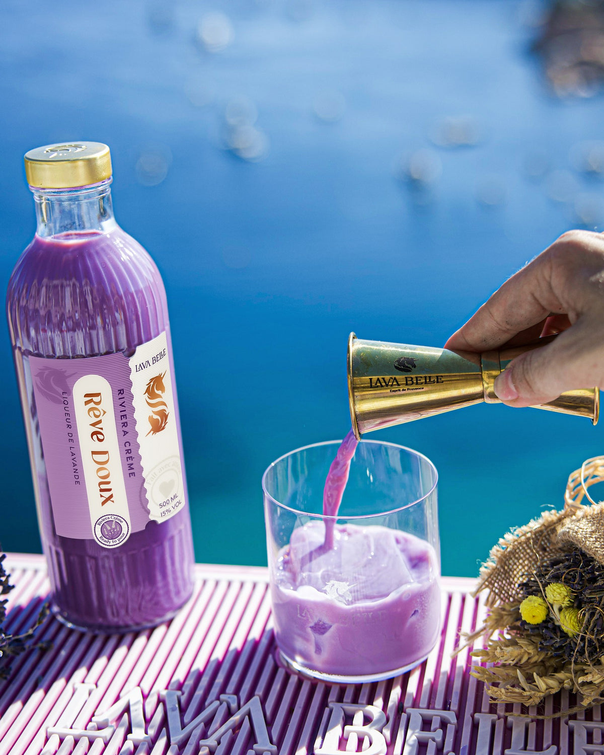 Cremiger violetter Rêve Doux wird aus dem Barmaß ins Glas gegossen, daneben die Flasche und ein Trockenblumenstrauß