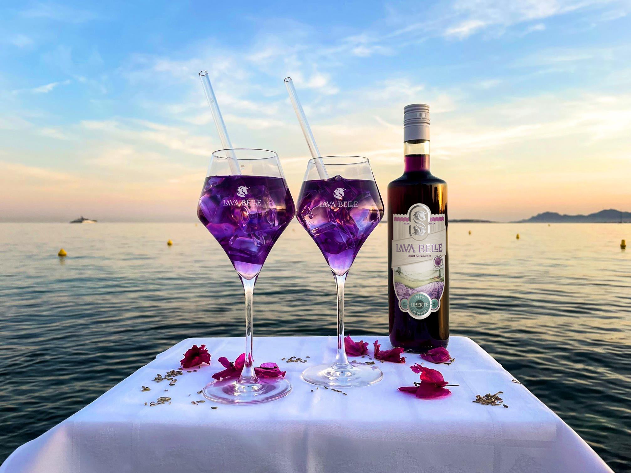 Zwei Gläser Lav'a Belle Spritz mit einer Flasche Liberté auf einem Tisch vor dem Hintergrund des ruhigen Wassers bei Sonnenuntergang am Strand von Antibes