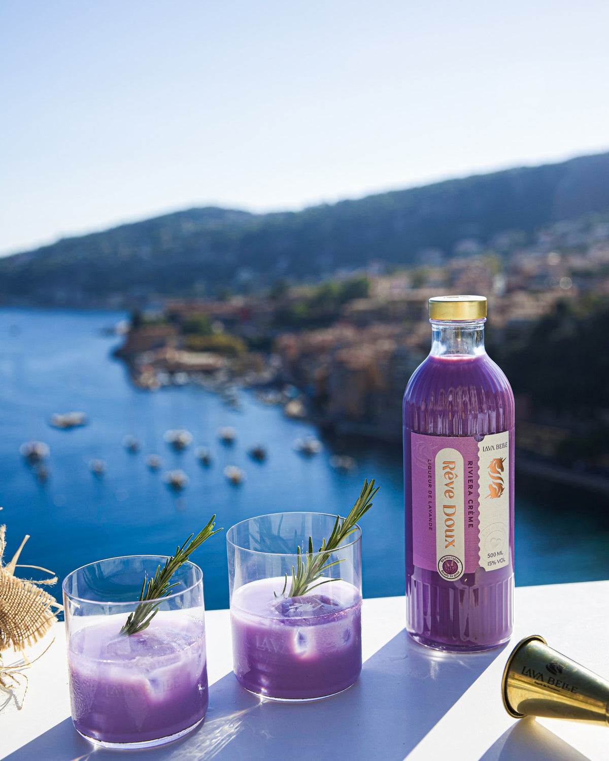 Flasche Lav'a Belle Rêve Doux und zwei Gläser mit cremig-violettem Drink und Rosmarin, dahinter eine Bucht an der Côte d'Azur