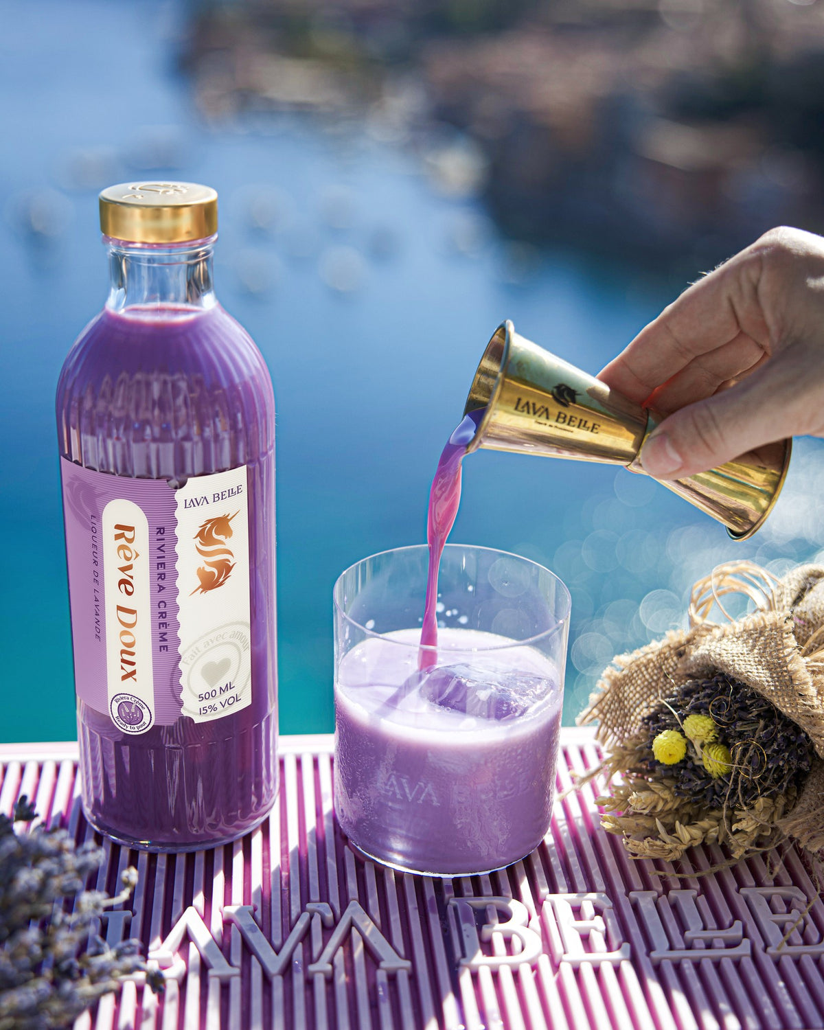 Rêve Doux wird aus dem goldenen Barmaß in ein Glas mit Eis gegossen, daneben die Flasche und getrockneter Lavendel