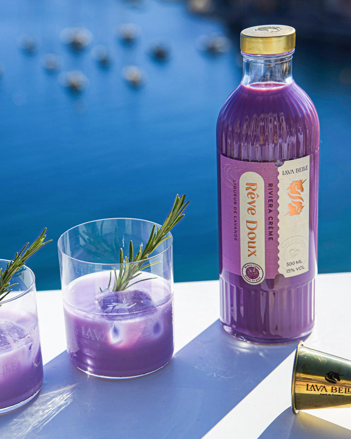 Flasche Rêve Doux neben zwei Gläsern mit Rosmarinzweig, im Hintergrund das Mittelmeer mit Booten